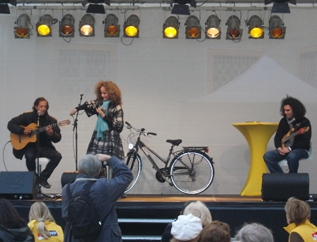 Sandra Pires und zwei Gitarristen auf einer Bühne, im Hintergrund ein Fahrrad.